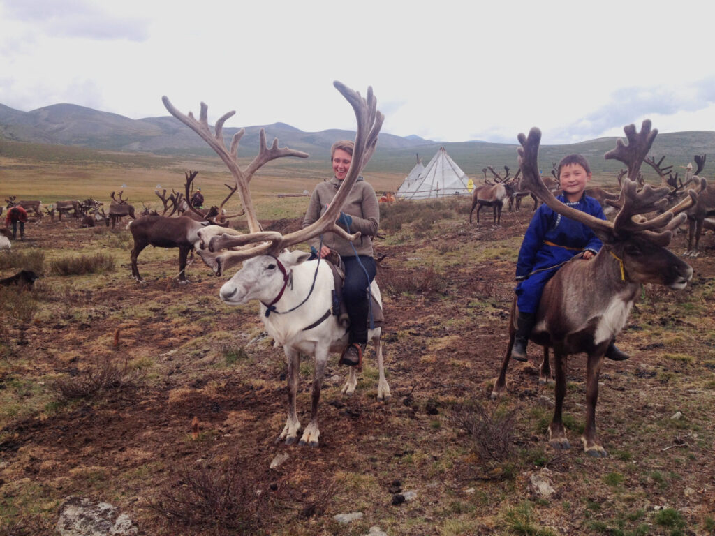 Suzanne on a reindeer next to a boy on a reindeer, on a mountain in Mongolia, both smiling.